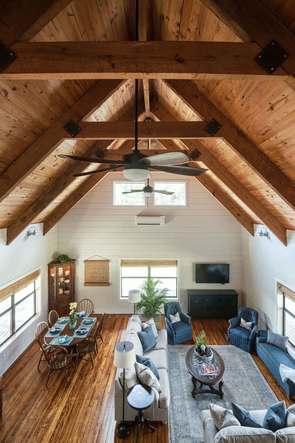 The living space features high ceilings with exposed wooden beams and large windows that allow natural light to fill the room. A spacious sofa and armchairs surround a coffee table, while a dining area with wooden chairs and a table is visible in the background.