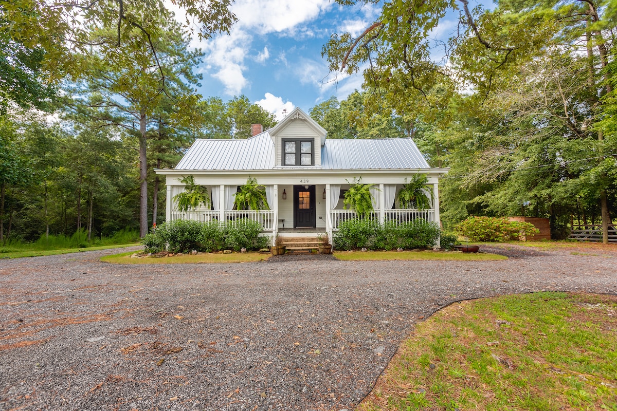 A charming farmhouse is nestled among trees, featuring a wraparound porch with white railing. The front door is framed by lush greenery, while a gravel pathway leads to the entrance. The structure's white exterior is complemented by a metal roof, blending rustic and modern elements.