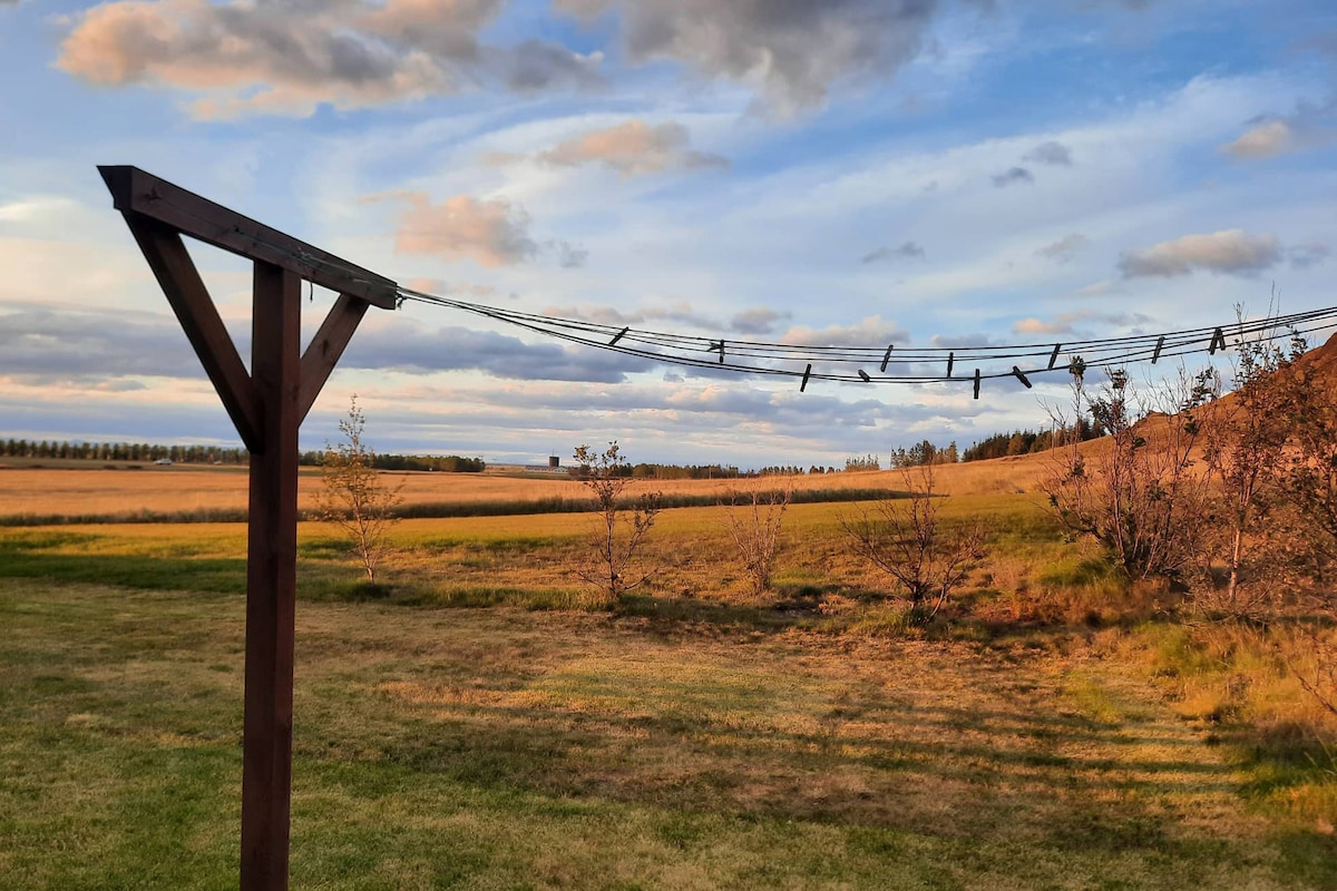 A clothesline is seen stretching between wooden posts in an open field, with greenery at the base. The landscape features gently rolling hills, under a sky with scattered clouds and soft evening light, reflecting the tranquility of the surroundings.