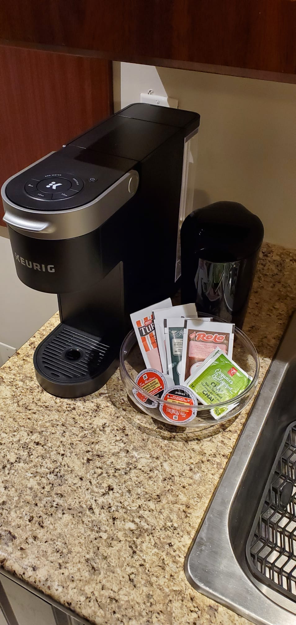 A coffee station is set up on a granite countertop, featuring a Keurig coffee maker alongside a bowl filled with assorted coffee pods and creamers. A small black container designed for used pods is also visible.
