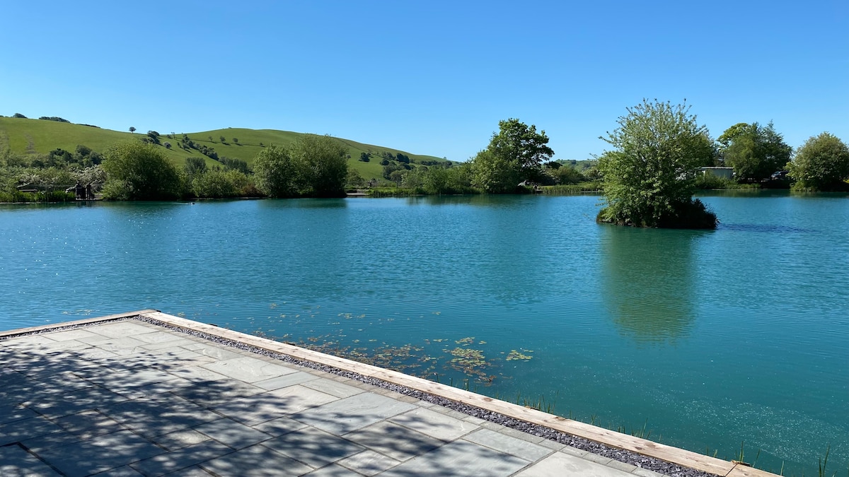 The image captures a serene lake surrounded by lush greenery and gentle hills. The clear water reflects the bright blue sky, with a small island visible in the center. A paved area leads to the water's edge, contributing to the tranquil atmosphere.