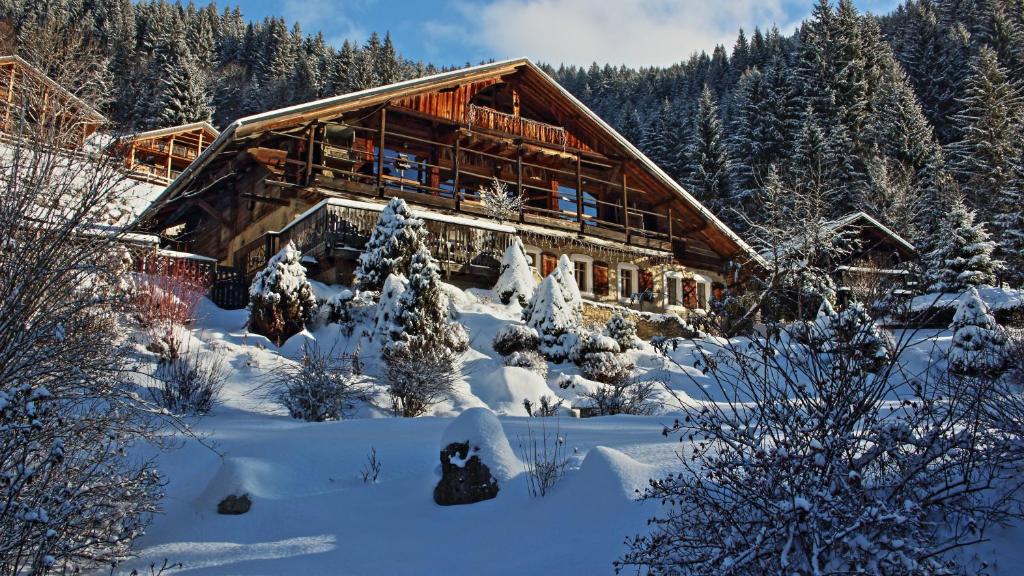 A charming alpine farmhouse is nestled amongst snow-covered greenery, surrounded by tall evergreen trees. The wooden structure features multiple balconies, which overlook the serene winter landscape. The foreground includes gently mounded snow interspersed with winter foliage, enhancing the tranquil setting.