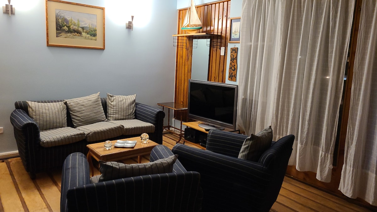 The living area features comfortable striped sofas arranged around a wooden coffee table. A television is positioned on a stand, complemented by soft lighting from wall sconces. Light streams in through sheer curtains that frame the door leading to the outdoor space.