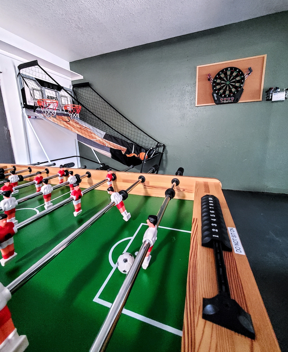 An entertainment area is showcased with a foosball table featuring red and white player figurines. Nearby, a basketball shooting game and a dart board are visible against the green wall, offering multiple options for recreation within the space.
