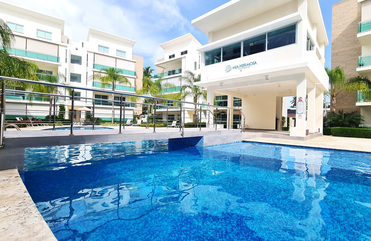 A clear blue swimming pool reflects the sky, surrounded by modern buildings with palm trees. The pool area is lined with smooth tiles, and a railing offers safety along the edge. Lounge chairs are available nearby for relaxation under the sun.