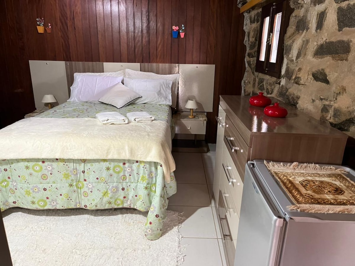 A cozy sleeping area is characterized by wooden paneling and stone accents. A spacious bed is covered with a floral-patterned blanket, complemented by white pillows. Nearby, a compact dresser and a small refrigerator are visible, along with decorative items on the dresser's surface.
