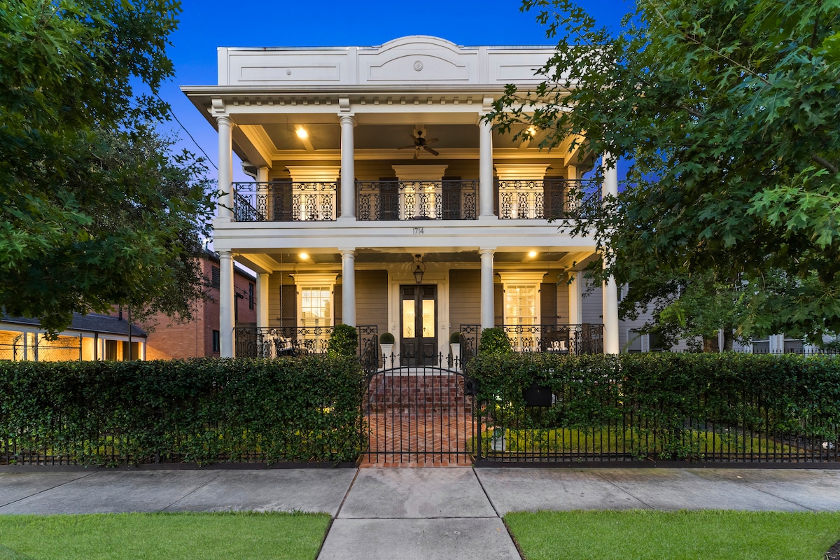 A classic two-story home is showcased, featuring large columns and intricate balconies. A well-maintained garden surrounds the property, with wrought iron fencing adding security. Warm lights illuminate the balconies and windows, creating an inviting evening ambiance.