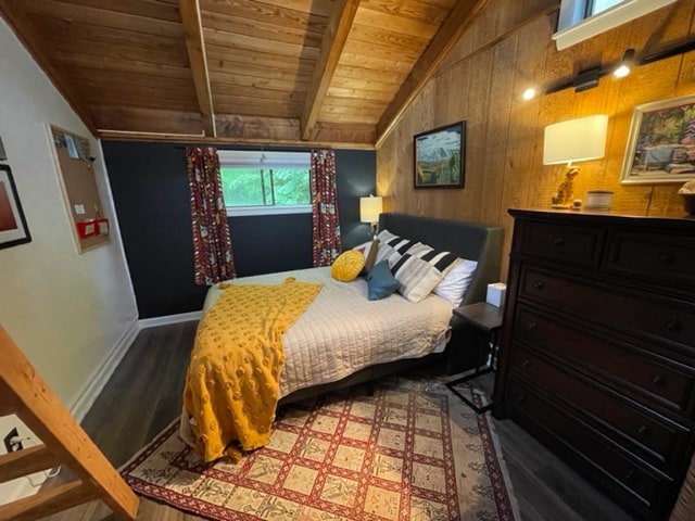 A cozy bedroom is highlighted by a queen bed adorned with patterned pillows and a golden throw. Natural wood beams support the ceiling, and a warm rug is positioned on the floor. A large window provides light, while a dresser and artwork add to the decor.