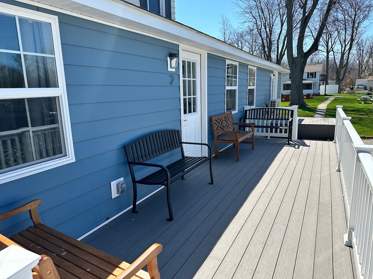 A covered porch area showcases a blend of seating options, including two black benches and a wooden bench, positioned on light gray composite decking. The entrance features a white door, with natural light filtering in through nearby windows, framed by the fresh blue hue of the exterior walls.