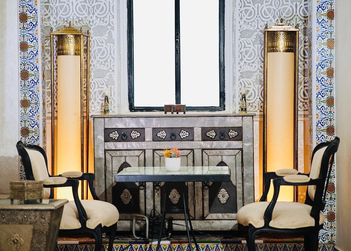 An elegant seating area is presented, featuring two dark wooden chairs with soft upholstery. A small table sits between them, topped with a potted plant. The backdrop showcases intricately patterned wall tiles and a stylish console, illuminated by warm light sources on either side.