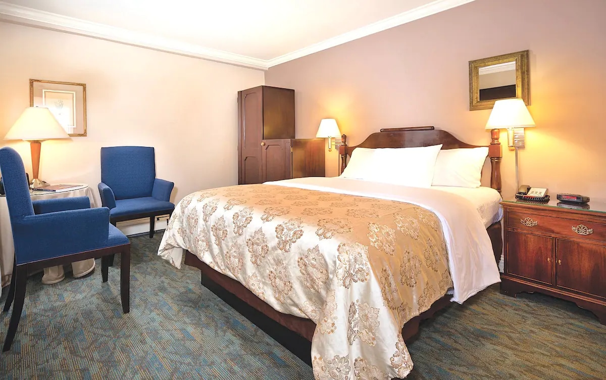 The room features a queen bed with a floral bedspread, complemented by two bedside lamps. A dark wood dresser is positioned beside the bed. A cozy armchair and a blue upholstered chair provide seating, and a decorative mirror adorns the wall.