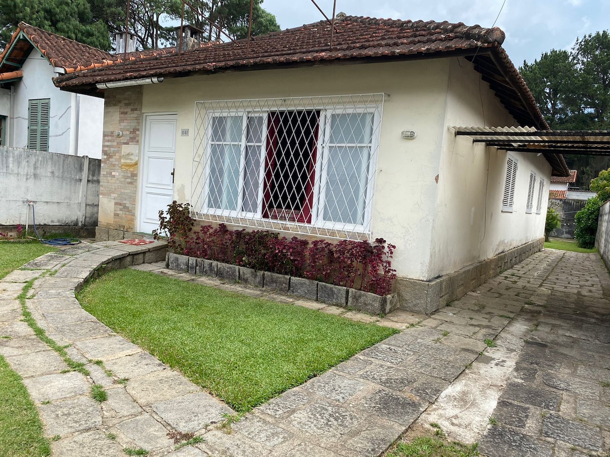 A single-story house is shown with a white exterior and a tiled roof. Decorative lattice covers the windows, which are framed by colorful plants. A stone pathway leads past a well-maintained lawn, providing access to the main entrance and side area.