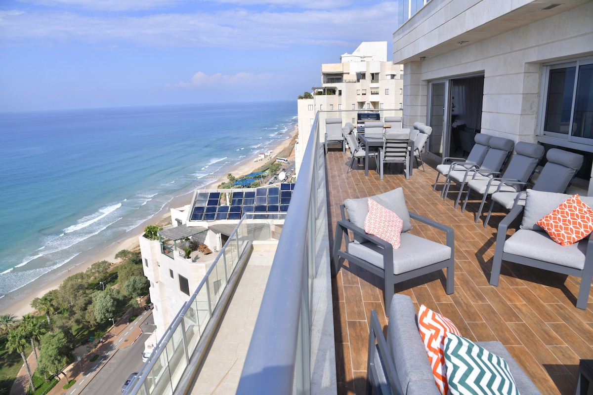 A spacious terrace offers stunning views of the coastline, with comfortable seating arrangements including lounge chairs. The area features decorative cushions and a polished wooden floor, creating an inviting outdoor space for relaxation and enjoyment.