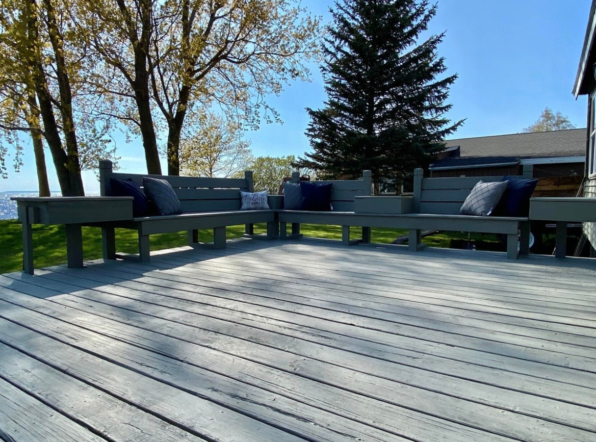 A spacious deck features several benches with cushions, positioned for comfort. Green grass surrounds the area, and a tall pine tree stands nearby. Lake Ontario is visible in the background under a clear blue sky, providing an inviting outdoor space for relaxation.