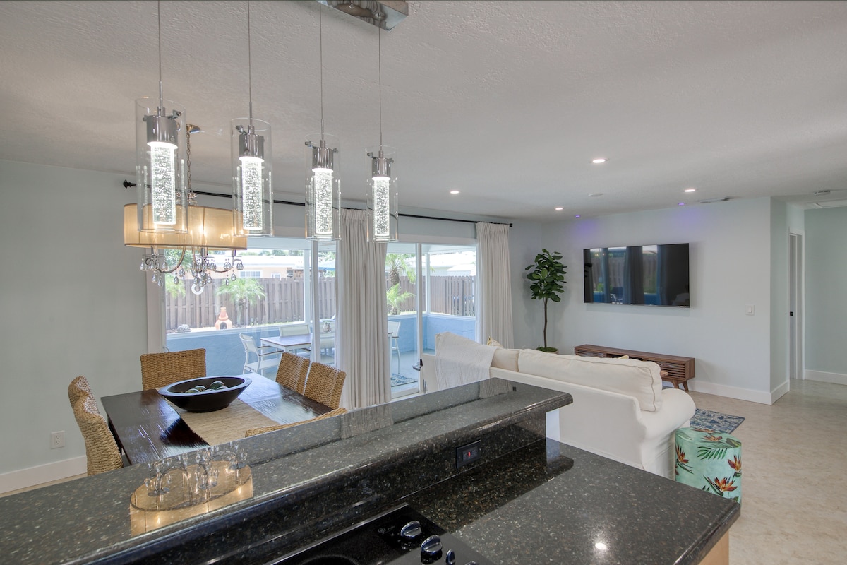 The open-plan dining and living area features a large dining table with woven seating for six. Overhead, modern light fixtures illuminate the space. A spacious sofa and a sleek entertainment unit with a flat-screen TV are visible, along with large windows offering natural light and views of the backyard.