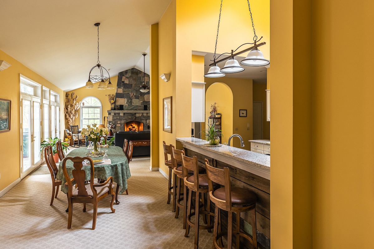 An open dining area features a long table adorned with a floral arrangement, surrounded by wooden chairs. A large stone fireplace is visible in the background, framed by bright yellow walls. Bar seating is provided at the kitchen counter, enhancing the communal space.