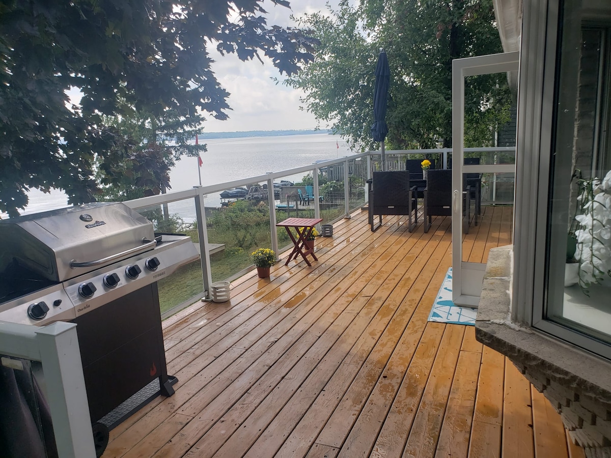 An outdoor deck area features a grilling station alongside seating arrangements. A small table is positioned near a potted plant. The expansive lake view is visible in the background, with boats anchored and greenery framing the scene.