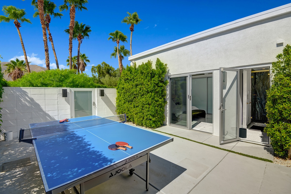 An outdoor area features a blue ping pong table positioned near lush greenery. Two sliding doors lead to a private courtyard, allowing for easy access. Palm trees are visible in the background against a clear blue sky, contributing to a vibrant and sunny outdoor space.