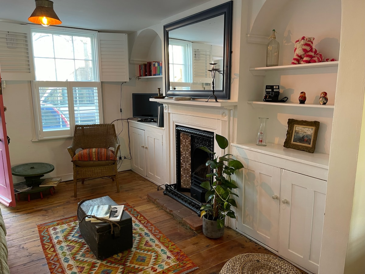 The open-plan living area features wooden flooring and a decorative fireplace. A comfortable armchair is positioned near a small table, with a television on a shelf nearby. Light enters through two windows with white shutters, enhancing the cozy ambiance.
