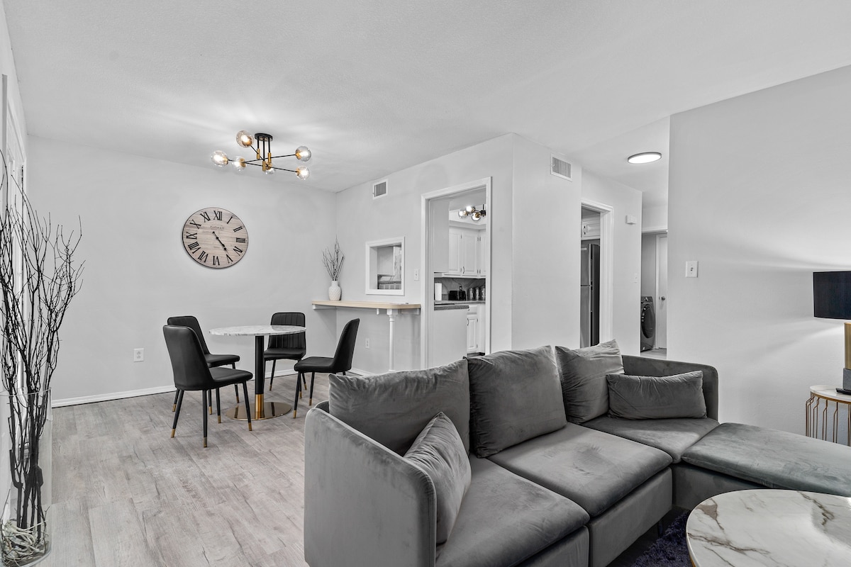 A modern living area features a plush gray sectional sofa and a circular dining table set for four. A wall clock hangs above, complementing the neutral tones of the space. Soft lighting creates an inviting ambiance throughout the open layout.