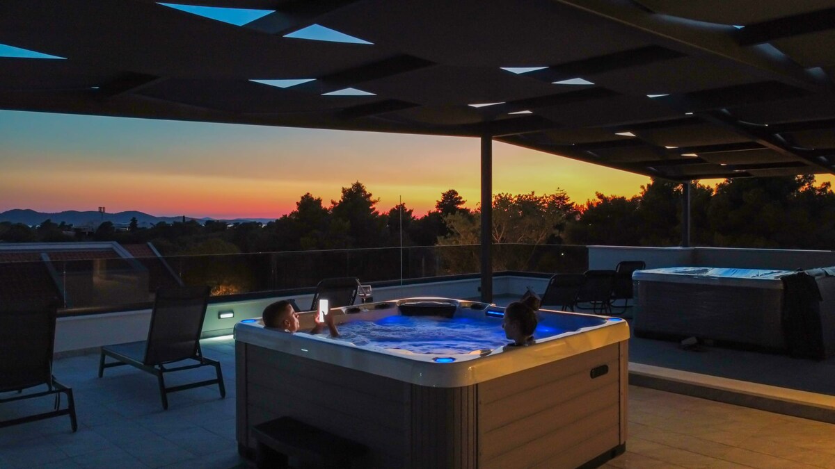 A spacious rooftop terrace is shown at sunset, featuring a jacuzzi for five people surrounded by lounge chairs. The warm colors of the setting sun illuminate the area, with a view of distant hills and trees in the background.