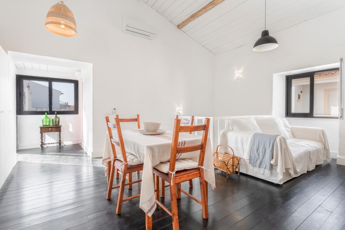 A bright open-plan living area features high vaulted ceilings, wooden floors, and large windows that allow natural light to fill the space. A dining table with wooden chairs sits beside a comfortable sofa bed, with a decorative basket placed nearby.