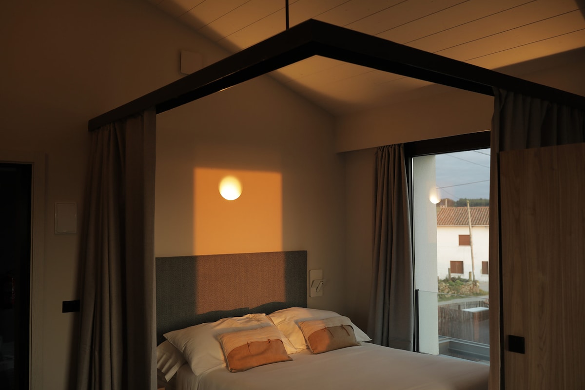 A modern bedroom features a cozy bed with light-colored linens accented by two decorative pillows. Soft light from a nearby window creates a warm ambiance, while curtains frame the space. A sleek pendant light is hung centrally above the bed.