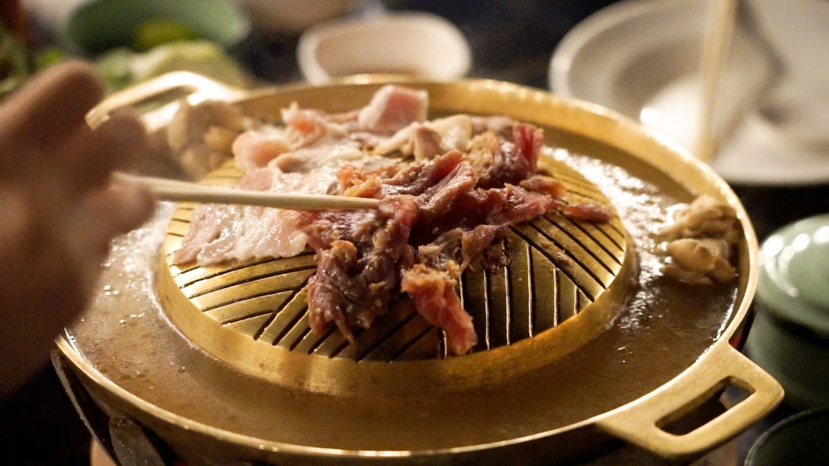 A hot grill is presented with thinly sliced meats, being cooked with gentle steam rising from the surface. Chopsticks are poised above the grill, ready to turn the cooking meat. An inviting array of fresh ingredients is visible in the background.