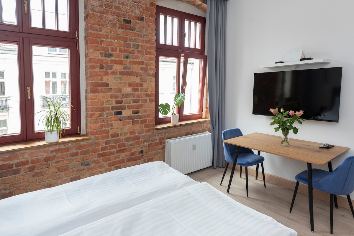 A bright room features exposed brick walls, enhancing the historic character of the space. Two large windows allow natural light to fill the area, complemented by a wooden table with two blue chairs. A flat-screen TV is mounted on the wall opposite the beds.