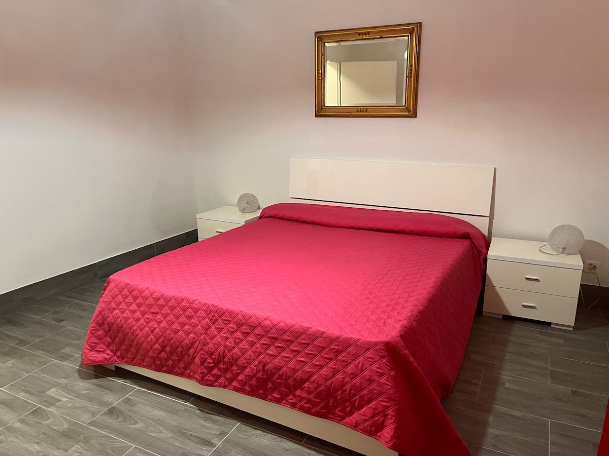 A bright bedroom features a spacious bed covered with a vibrant red quilt. Two bedside tables with lamps are positioned on either side of the bed. A large mirror hangs above the headboard, reflecting the simple elegance of the room.