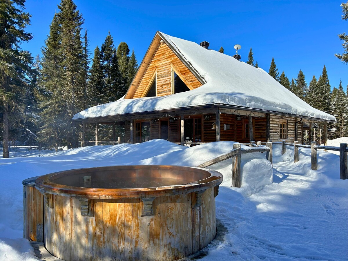 A wooden chalet with an A-frame structure sits amidst a snow-covered landscape, surrounded by tall coniferous trees. A circular hot tub is positioned in the foreground, with steam rising gently into the crisp air, enhancing the serene winter setting.