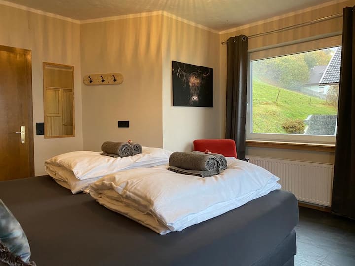 Двойната спалня с легло KingSize Boxspring, фотьойл, монтирано огледало и закачалки за гардероб.