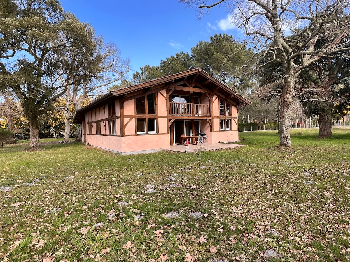 The exterior of the house features a wooden structure with large windows and a balcony. A spacious lawn surrounds the home, adorned with trees and natural greenery, creating a peaceful setting. The facade is characterized by a soft earth-tone color, complementing the tranquil environment.