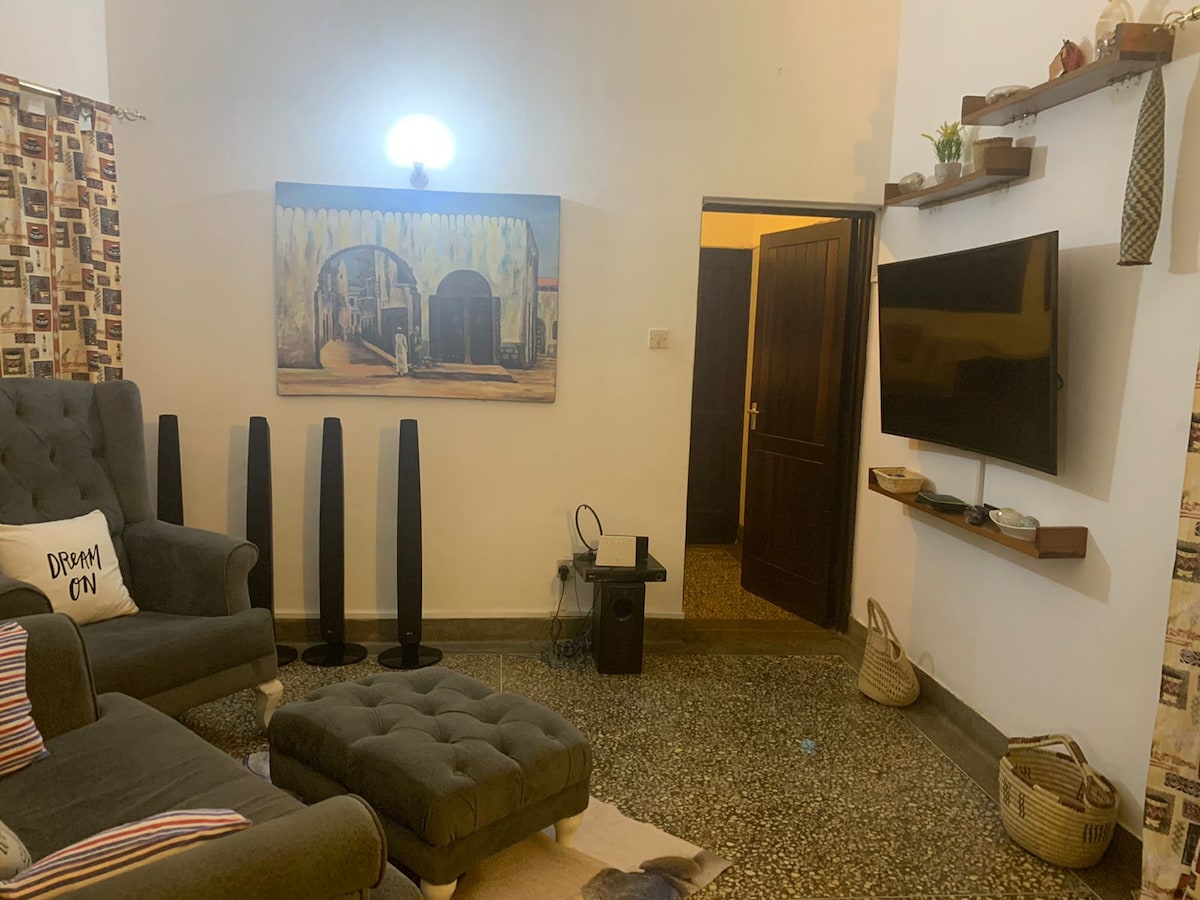 A living area features a plush gray couch with colorful throw pillows. A large wall art piece depicting an archway hangs above a sleek TV mounted on the wall. Two tall, slender speakers stand beside the couch, enhancing the room's modern aesthetic.