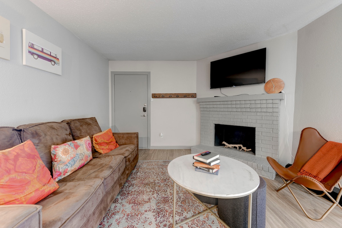 A living room area features a comfortable sofa adorned with decorative pillows, facing a flat-screen TV mounted above a brick fireplace. A round coffee table sits in the center, accompanied by a cozy accent chair. Light streaming in enhances the neutral tones of the space.