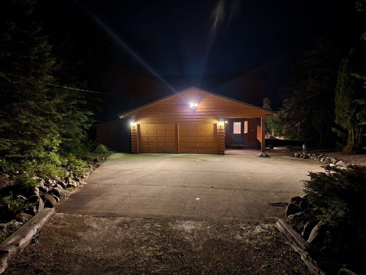 A welcoming wooden exterior is illuminated by warm lighting, showcasing a spacious driveway leading to a two-car garage. Surrounding trees provide privacy, hinting at a serene atmosphere. The entrance of the home offers a glimpse of light from the interior.