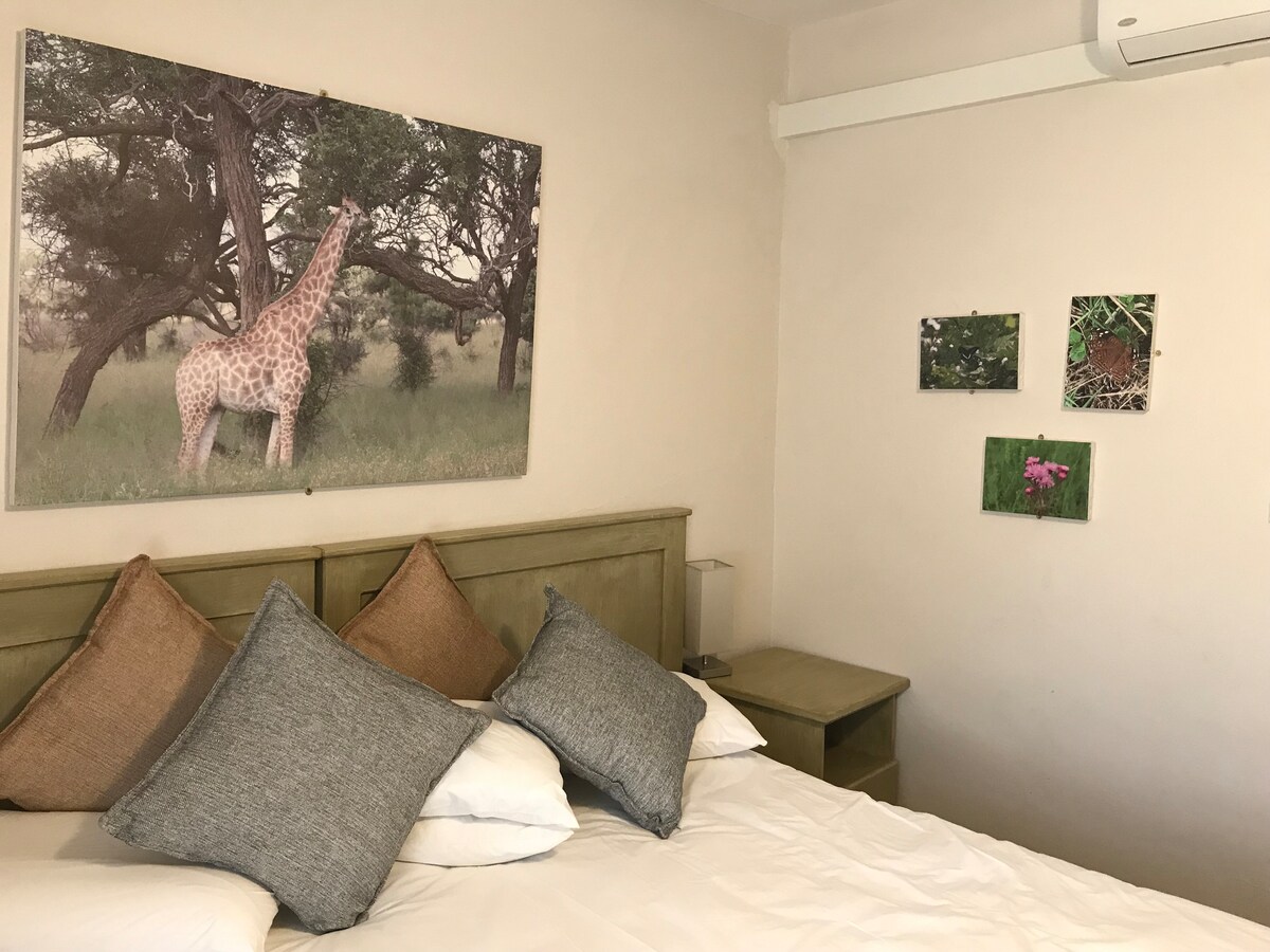 A spacious bed is adorned with several decorative pillows, positioned against a subtle wall. Above the bed, a large photograph of a giraffe in a natural setting adds visual interest. Nearby, smaller framed images of plants and flowers enhance the decor.