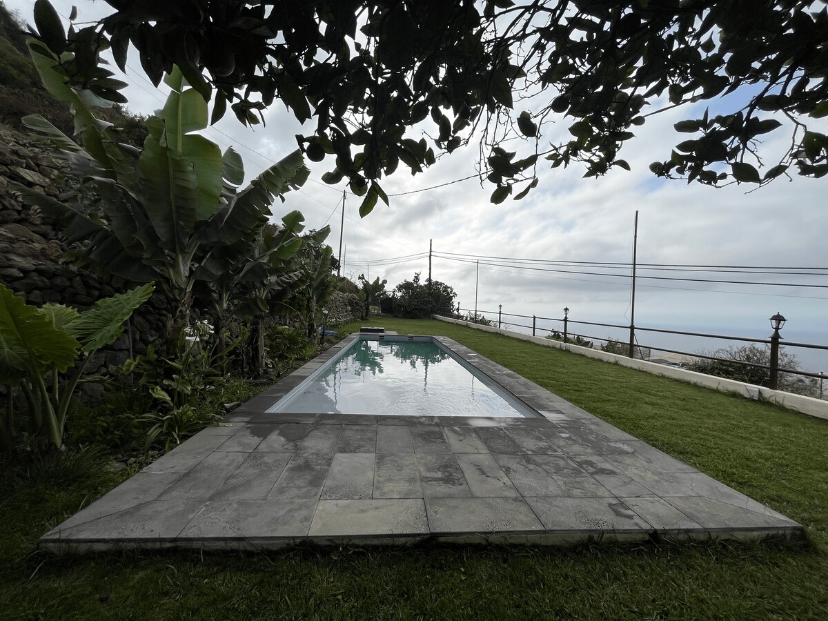 A serene saltwater pool is surrounded by well-maintained grassy areas and tropical plants. The pool's surface reflects the sky and nearby landscape, creating a tranquil setting. Stone tiles provide an even patio around the pool, enhancing the natural beauty of the surroundings.