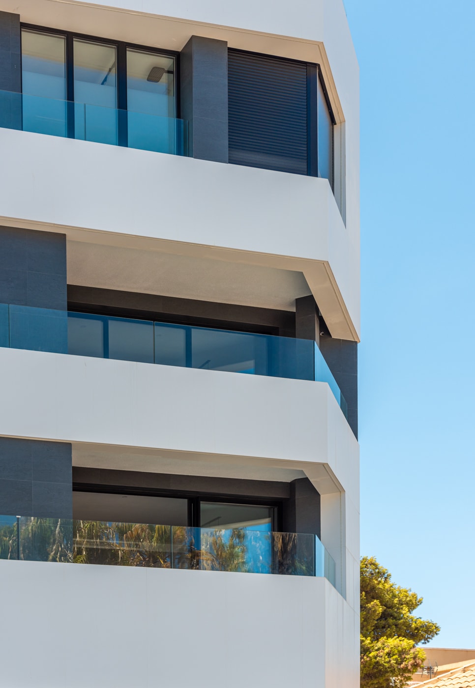 A modern apartment building is depicted, featuring clean lines and a combination of white and gray facades. Large glass balconies provide unobstructed views, while greenery is visible at the base, enhancing the contemporary architectural design.