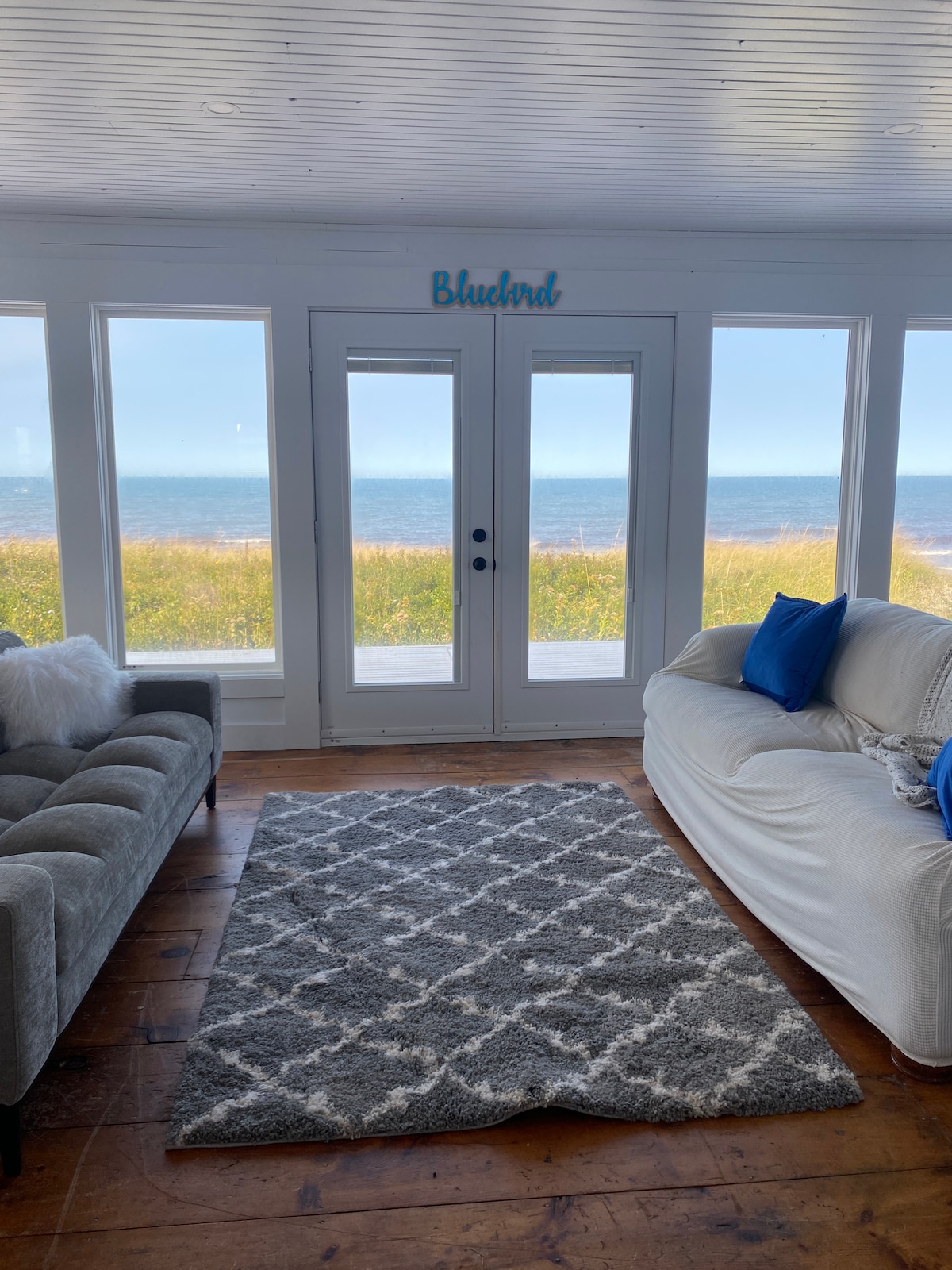 An open living area is surrounded by large windows, providing views of the ocean and grassy landscape outside. A grey rug covers the hardwood floor, with a light-colored sofa and a plush grey couch positioned to face the view.