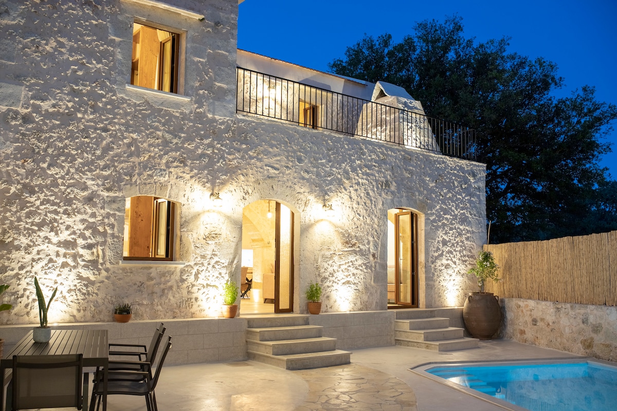 The exterior of the villa is highlighted by a rustic stone façade, softly illuminated by outdoor lighting. Large arched windows allow glimpses into the interior, while steps lead down to a private pool area surrounded by a paved patio and landscaped greenery.