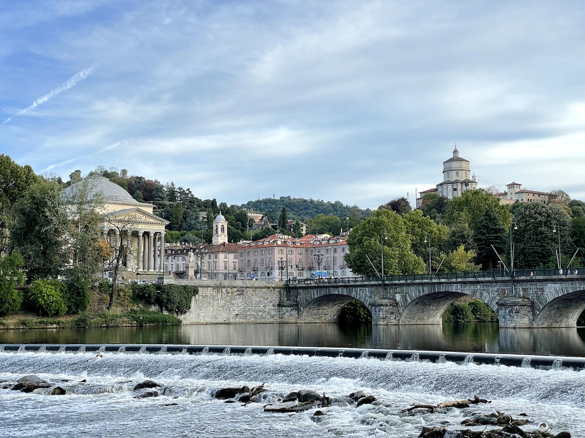 A serene view of a river with gentle rapids, framed by lush greenery. In the background, historic buildings, including a dome-shaped structure, are positioned along the riverbank, showcasing architectural details. A stone bridge spans the river, connecting the two sides of the scene.