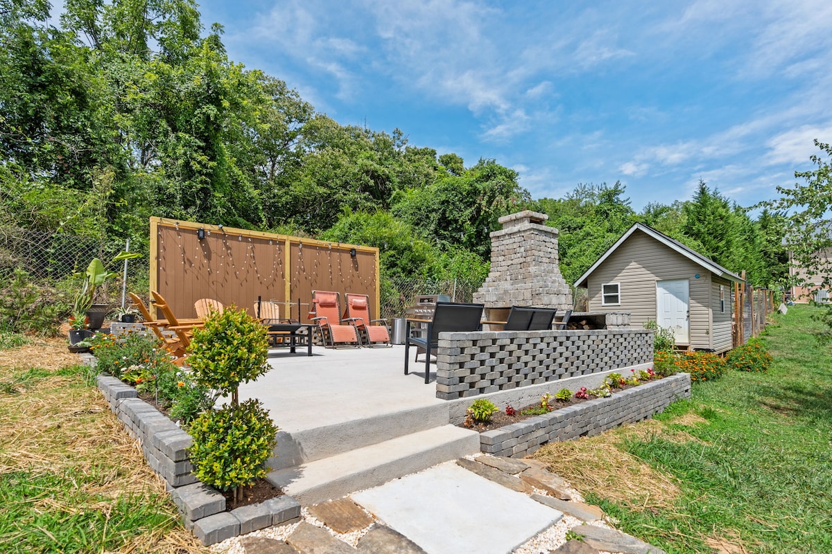 An outdoor space features a concrete patio with a stone fireplace at its center. Surrounding seating includes wooden chairs and lounge chairs, with a backdrop of greenery. A wooden wall adorned with string lights adds a cozy touch to the gathering area.