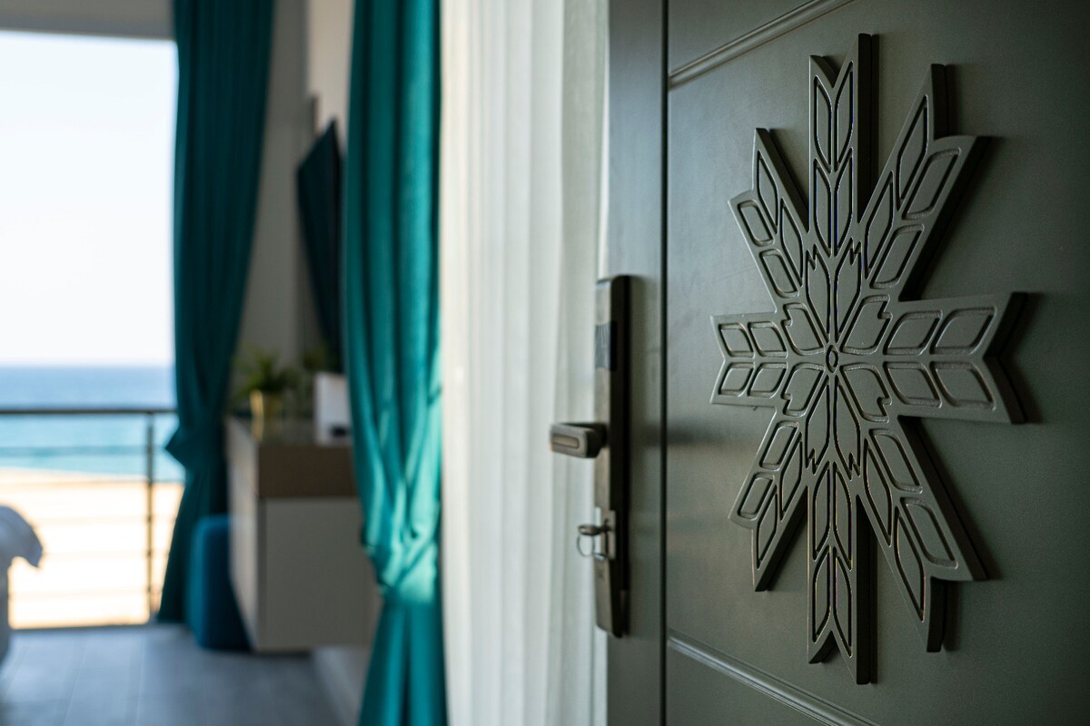A decorative door featuring an intricate snowflake design is in focus, framed by sheer curtains and vibrant turquoise drapes. A glimpse of the ocean view is visible through the doorway, highlighting the serene coastal atmosphere.