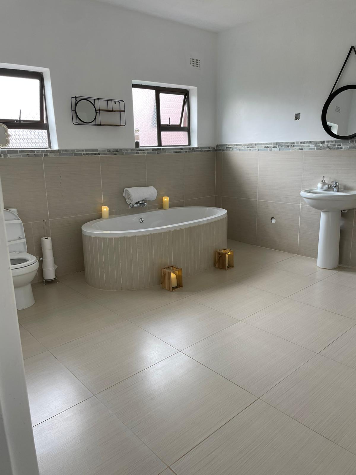 A spacious bathroom features a freestanding oval bathtub, complemented by two windows that provide natural light. A pedestal sink and toilet are present, with soft lighting from decorative lanterns placed on the floor. Neutral tiles create a calm and clean atmosphere.