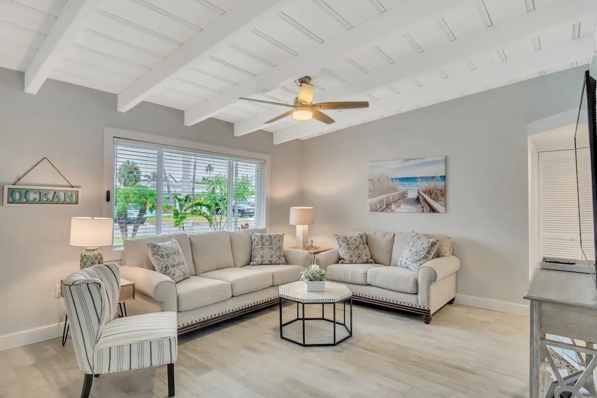 A spacious living area is highlighted by a sectional sofa and a modern coffee table. Natural light floods through the windows, revealing a serene view of outdoor greenery. A ceiling fan adds a cool breeze, and soft lamps provide cozy lighting for relaxation.