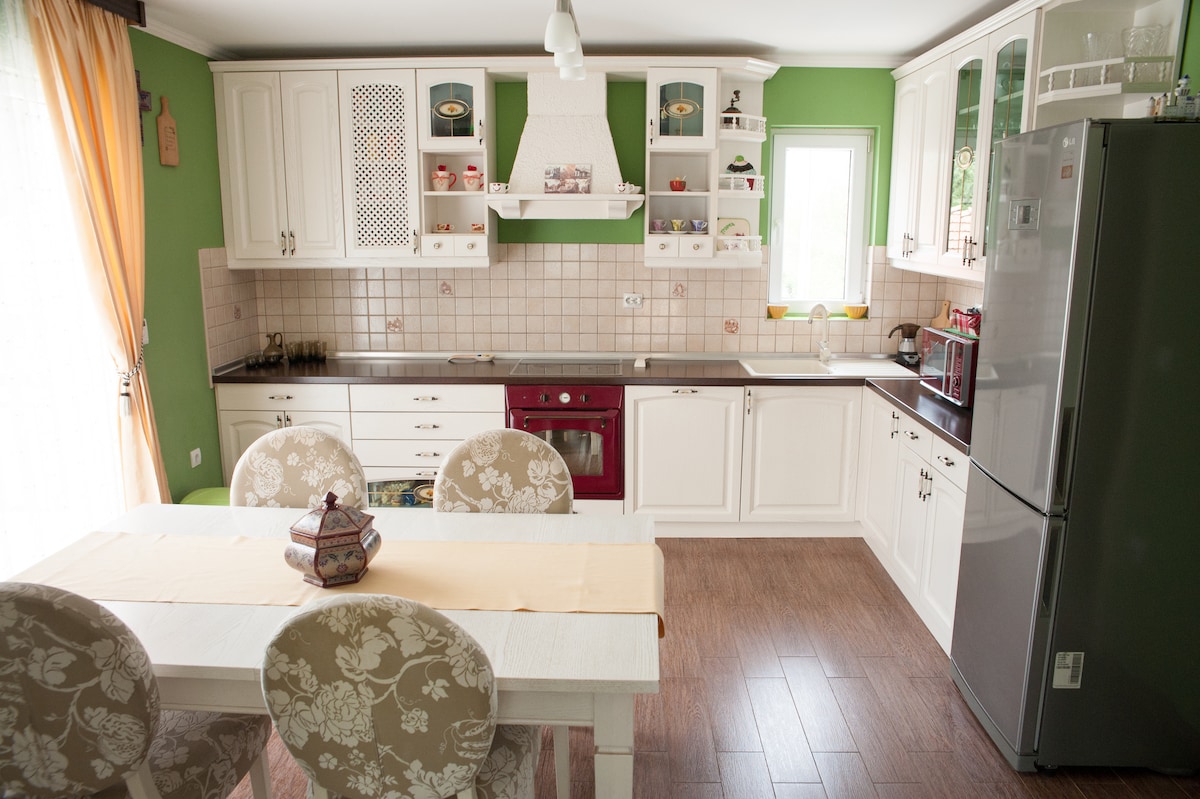 A well-equipped kitchen is presented, featuring light cabinetry with an open design. A dining table with floral-patterned chairs is positioned centrally. Modern appliances, including a refrigerator and stove, complete the space. Natural light is allowed in through the window, enhancing the overall function of the area.