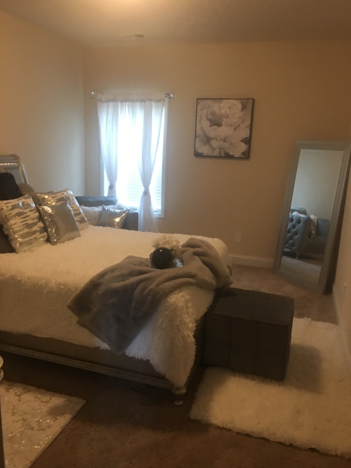A tranquil bedroom features a spacious bed adorned with decorative pillows and a soft throw blanket. Natural light filters through a window with sheer curtains. A tall mirror and a plush ottoman enhance the room's comfort, complemented by a cozy area rug.
