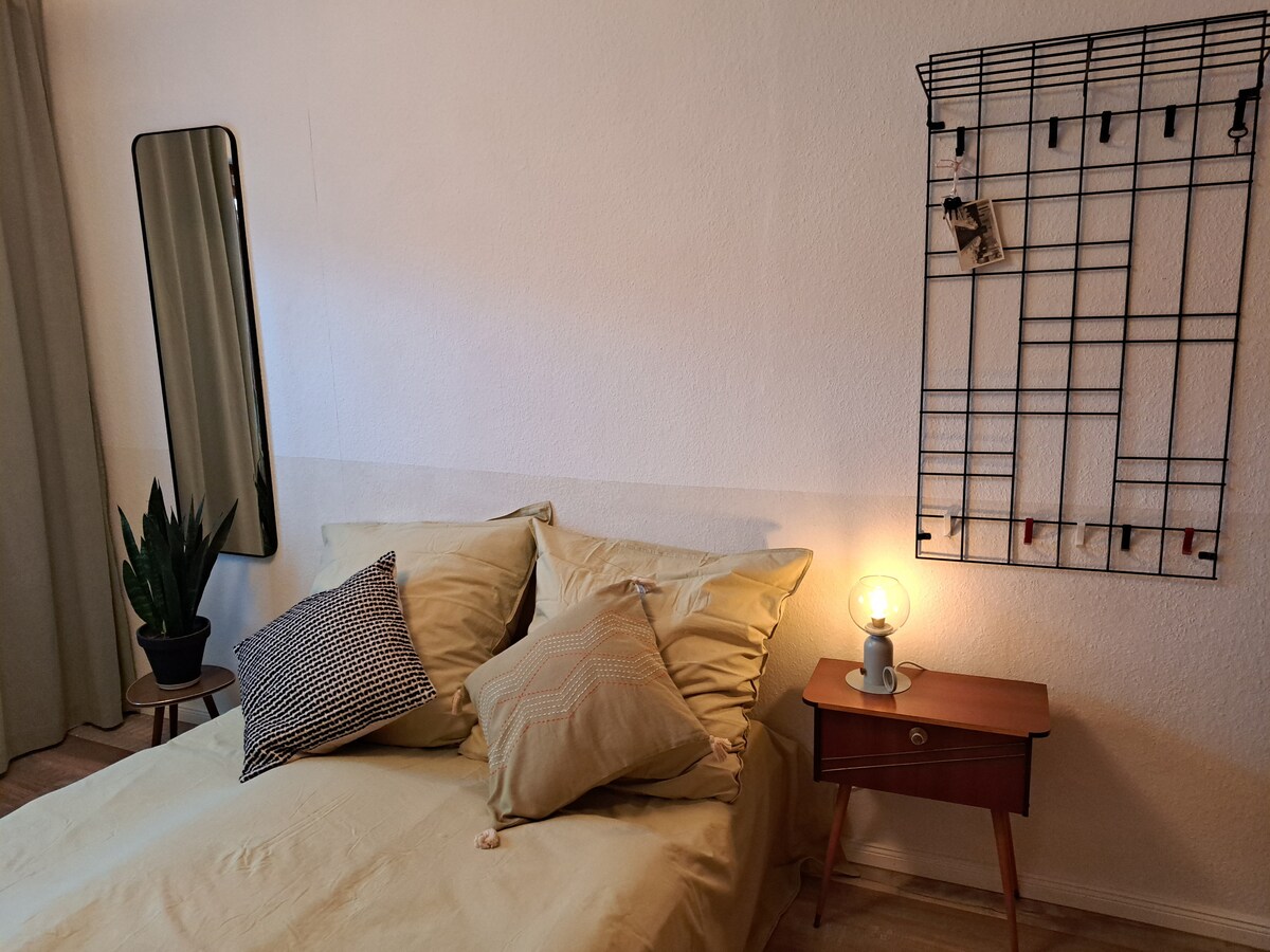 A calming bedroom is featured, highlighting a spacious bed adorned with soft pillows and a muted bedspread. A warm bedside lamp and a wooden nightstand add functionality. A large mirror reflects light, while a plant in the corner introduces a touch of greenery.