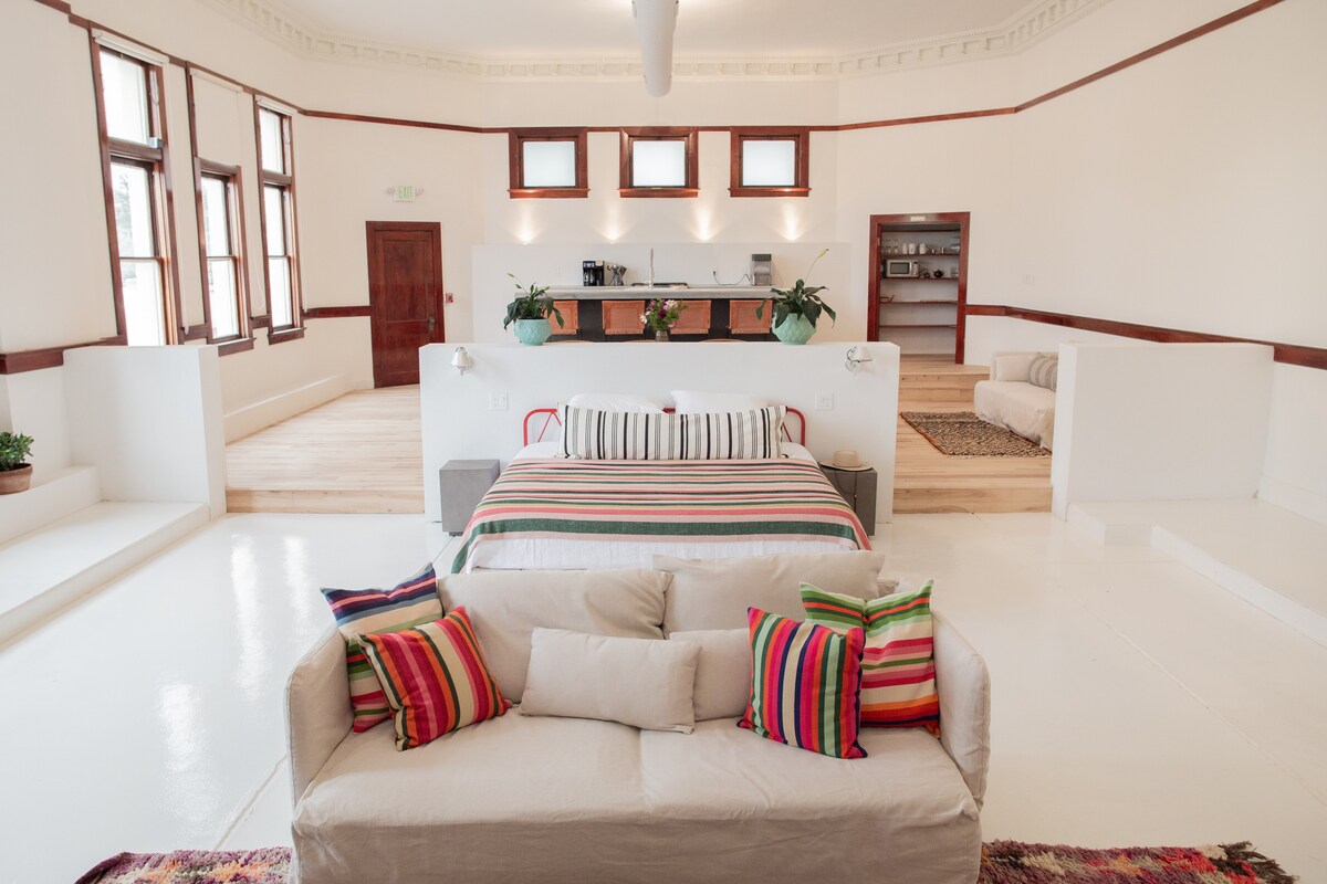 An open living space features a spacious bed adorned with striped bedding, framed by a light-colored sofa decorated with colorful cushions. Bright windows allow natural light to fill the room, while a modern kitchen area is visible in the background.